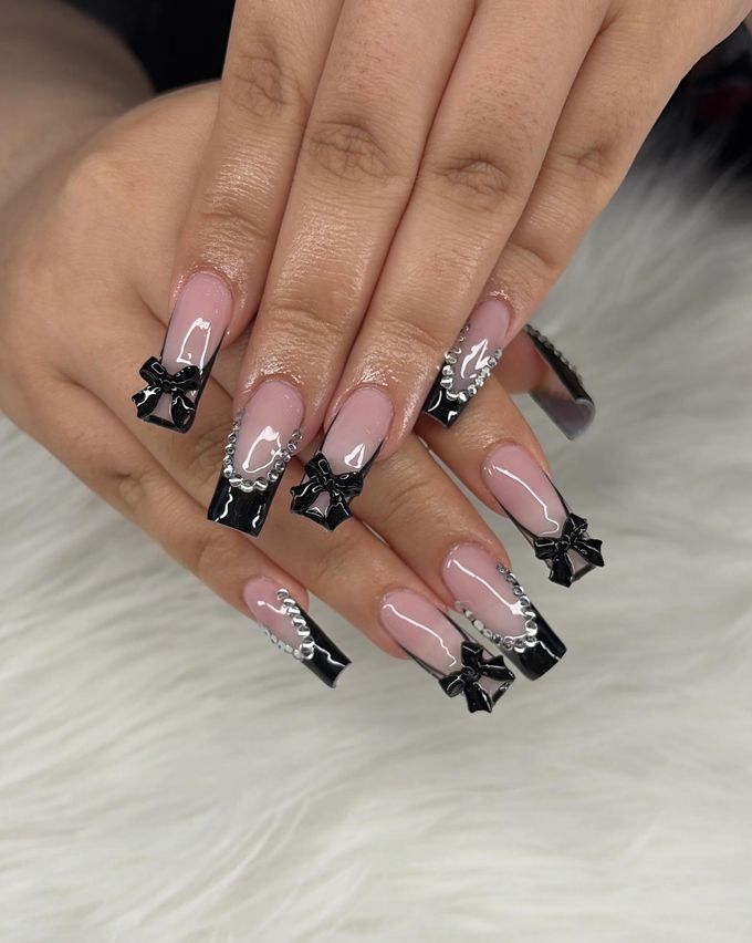 Long coffin Gel X nails with sheer pink bases, glossy black French tips, raised black 3D bows and arcs of small rhinestones, resting on white fur.